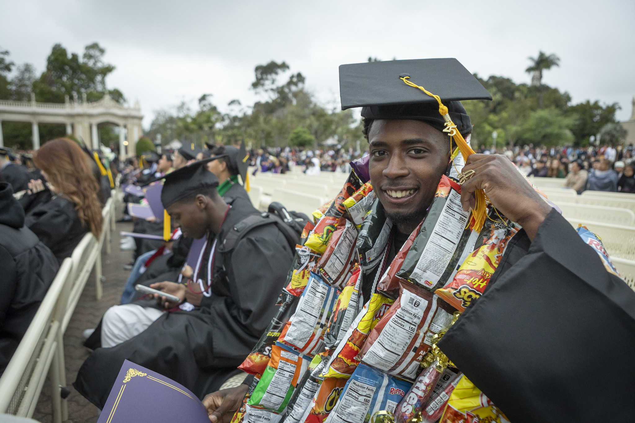
A graduate wearing a necklace made out of about 20 small bags of Cheetos seated in the crowd
