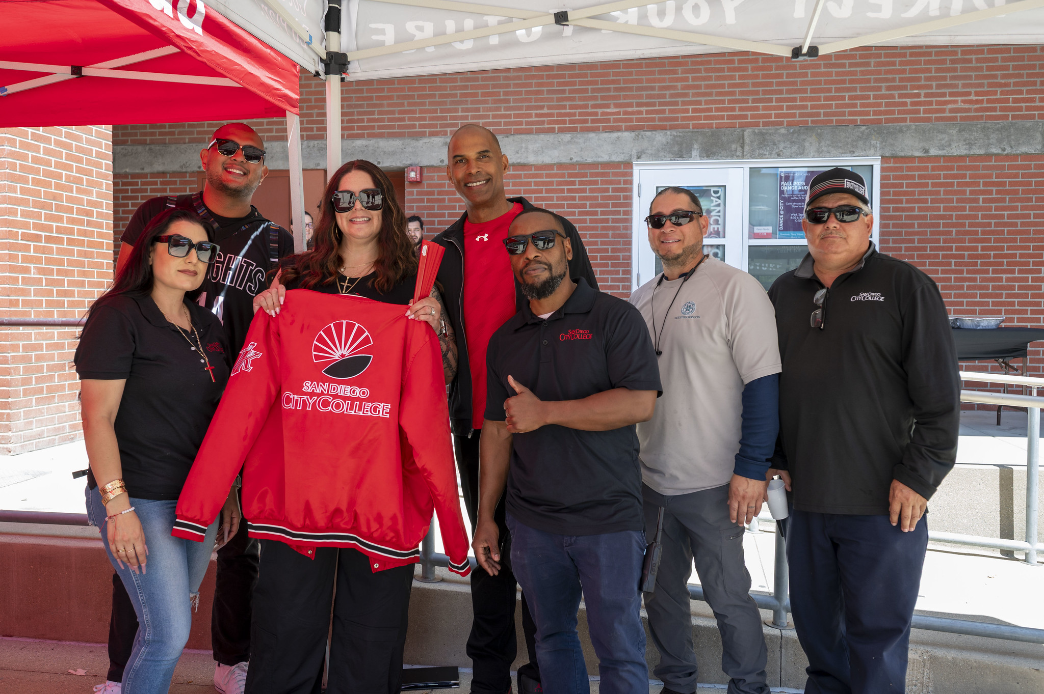
Seven employees under a tent at Convocation. One is holding up a red City College jacket.
