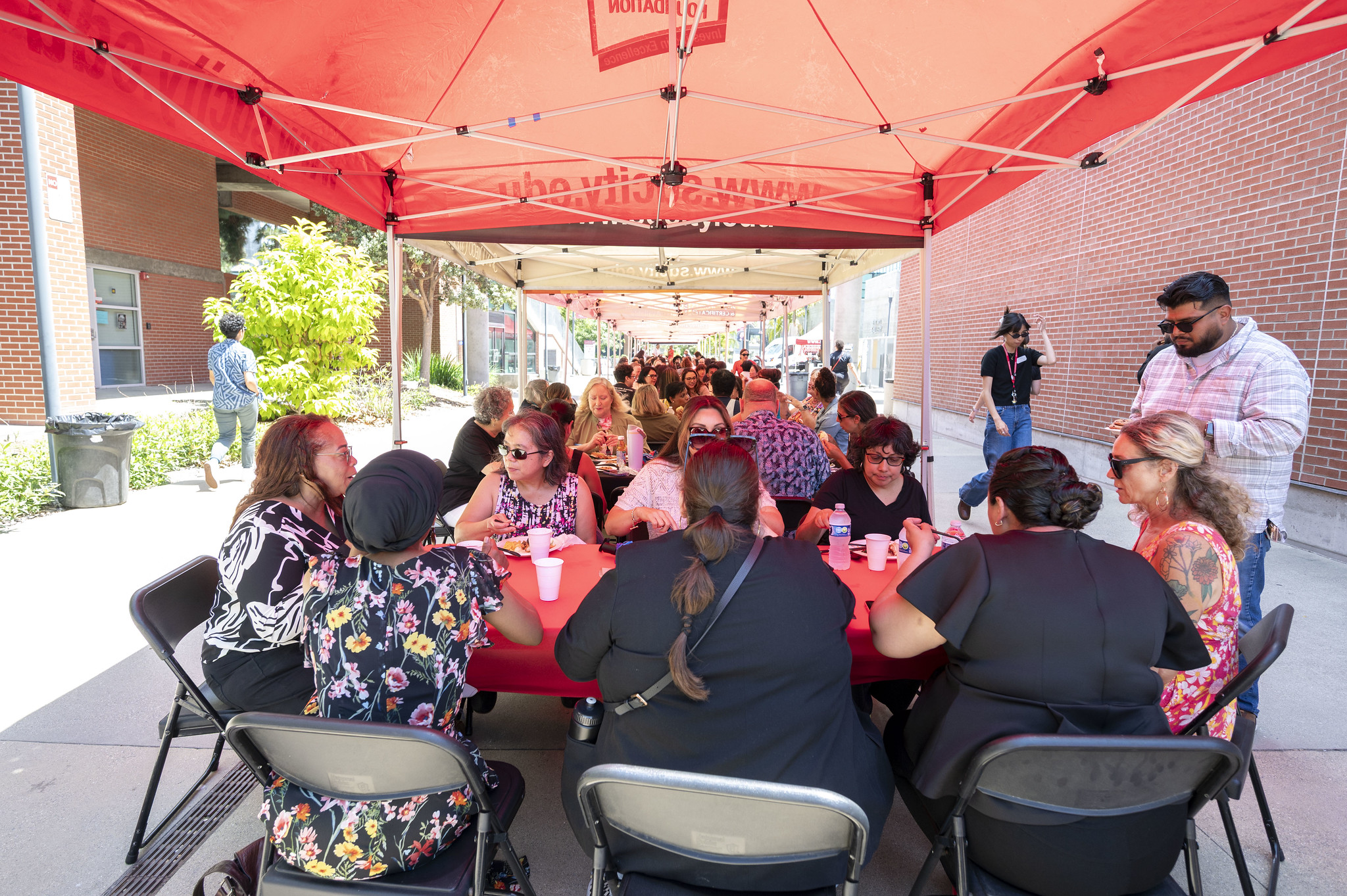 
Tables of people have lunch under red City College tents.
