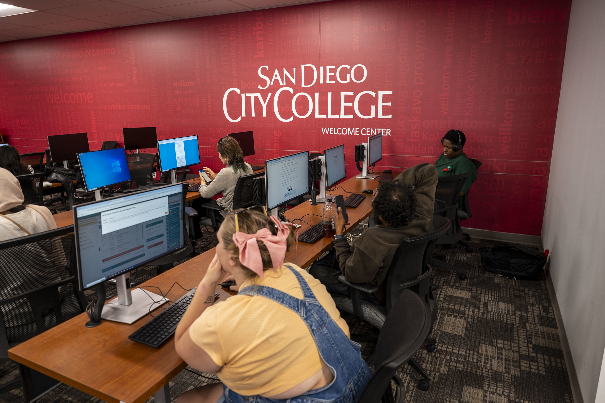 
A classroom full of people sit at computer stations. The wall on their right is red with the City College logo.
