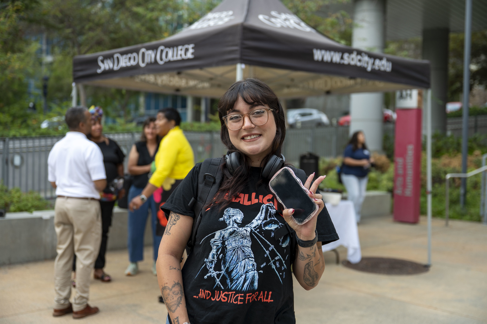 
A student near a welcome tent wearing headphones around her neck and holding her phone waves to the camera.
