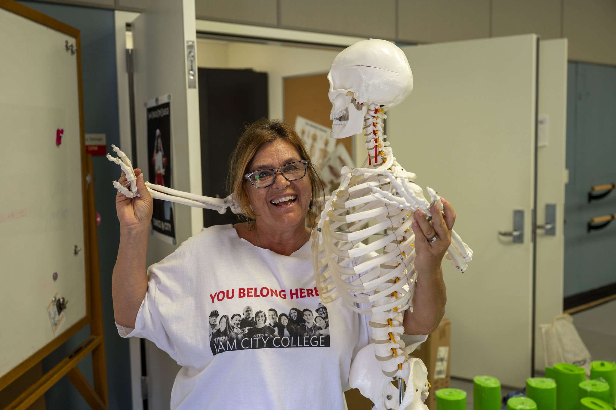 
An instructor in a classroom is smiling with her arm around a skeleton.
