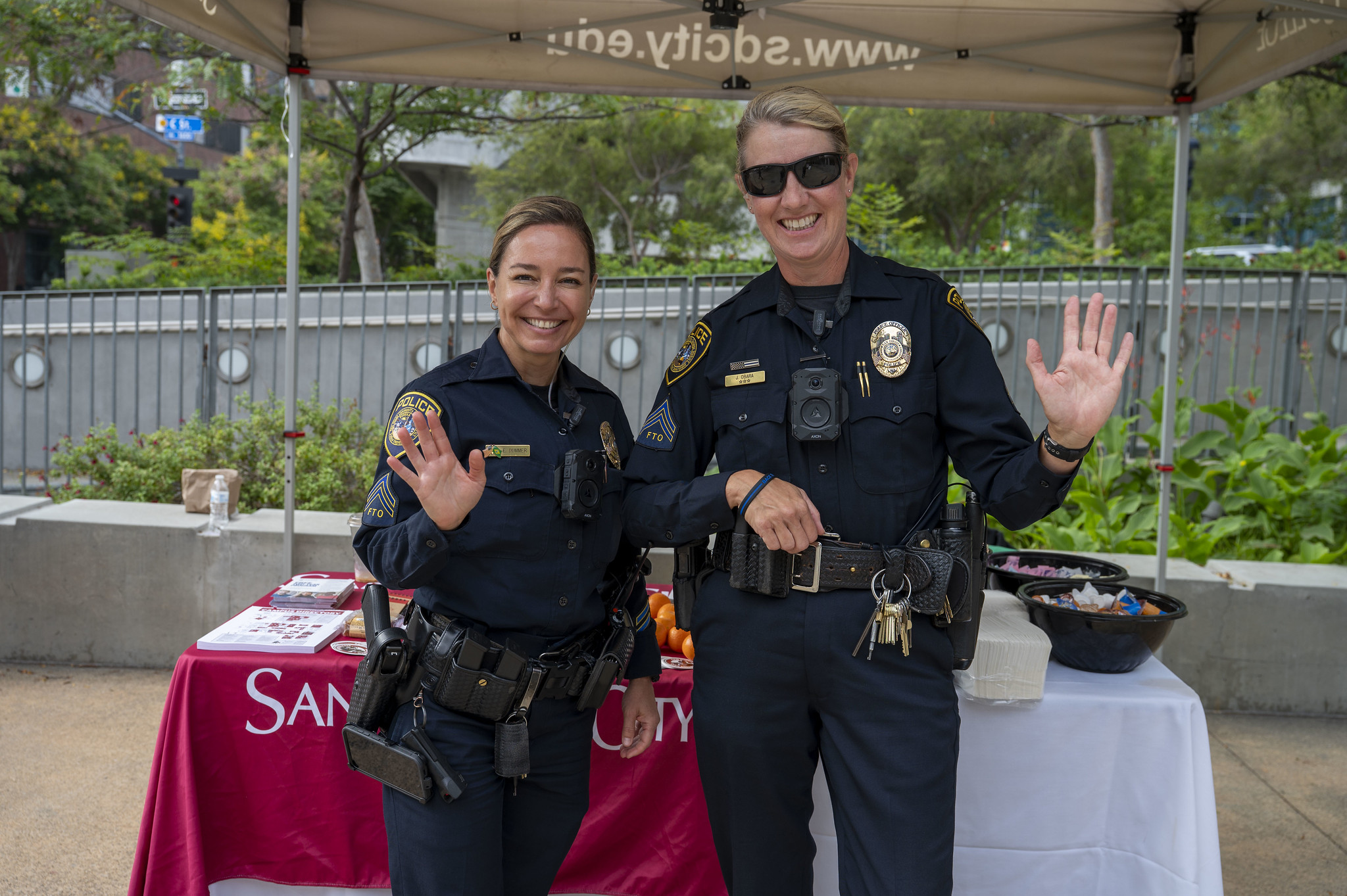 
Two college police officers smile and wave to the camera.&nbsp;
