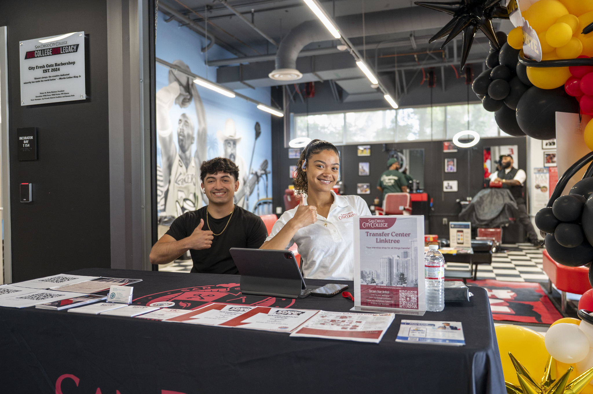 
Two people working a transfer table outside of Fresh Cutz Barbershop give a thumbs up
