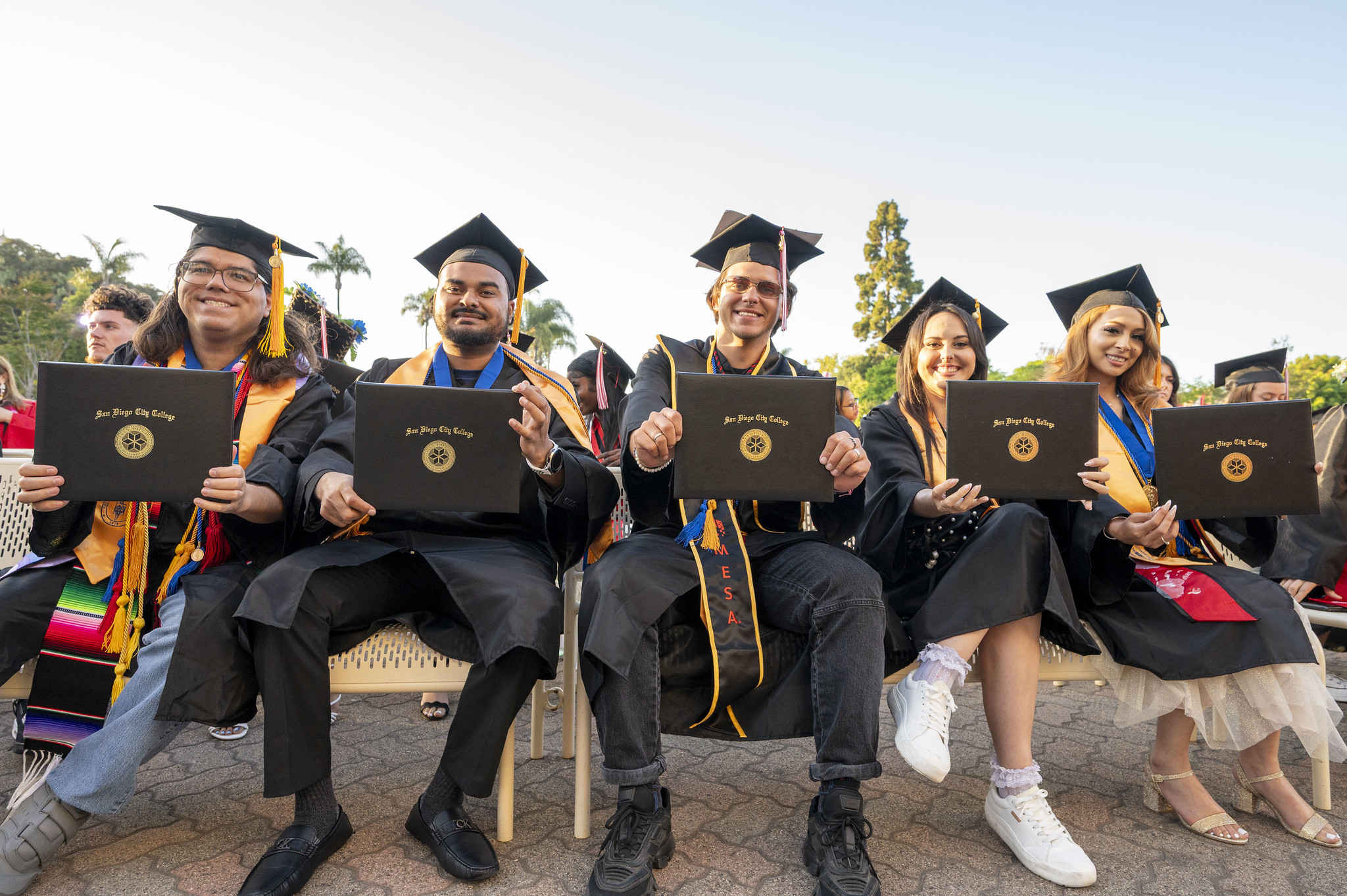 
Five graduates seated on a bench holding up their degrees.
