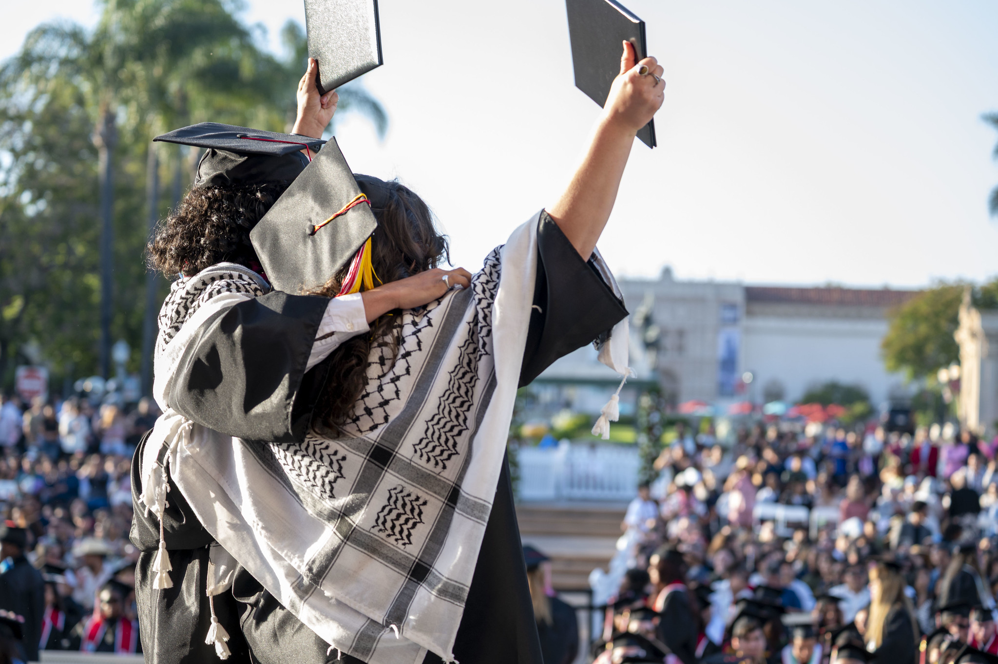 
Two graduates with their arm around each other hold up their degrees to the audience.
