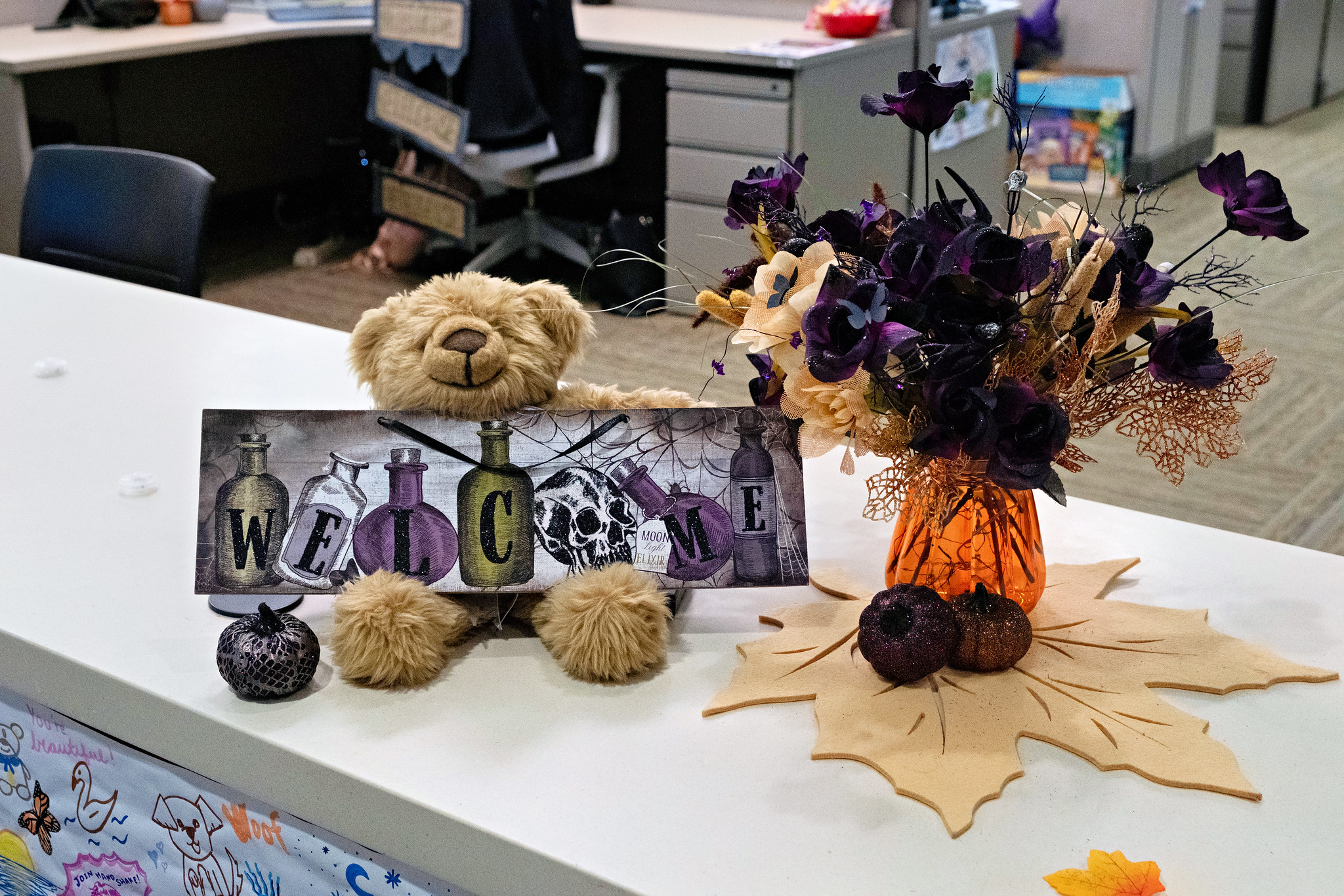 
A Teddy bear on a counter with a welcome sign seated next to some fall flowers.
