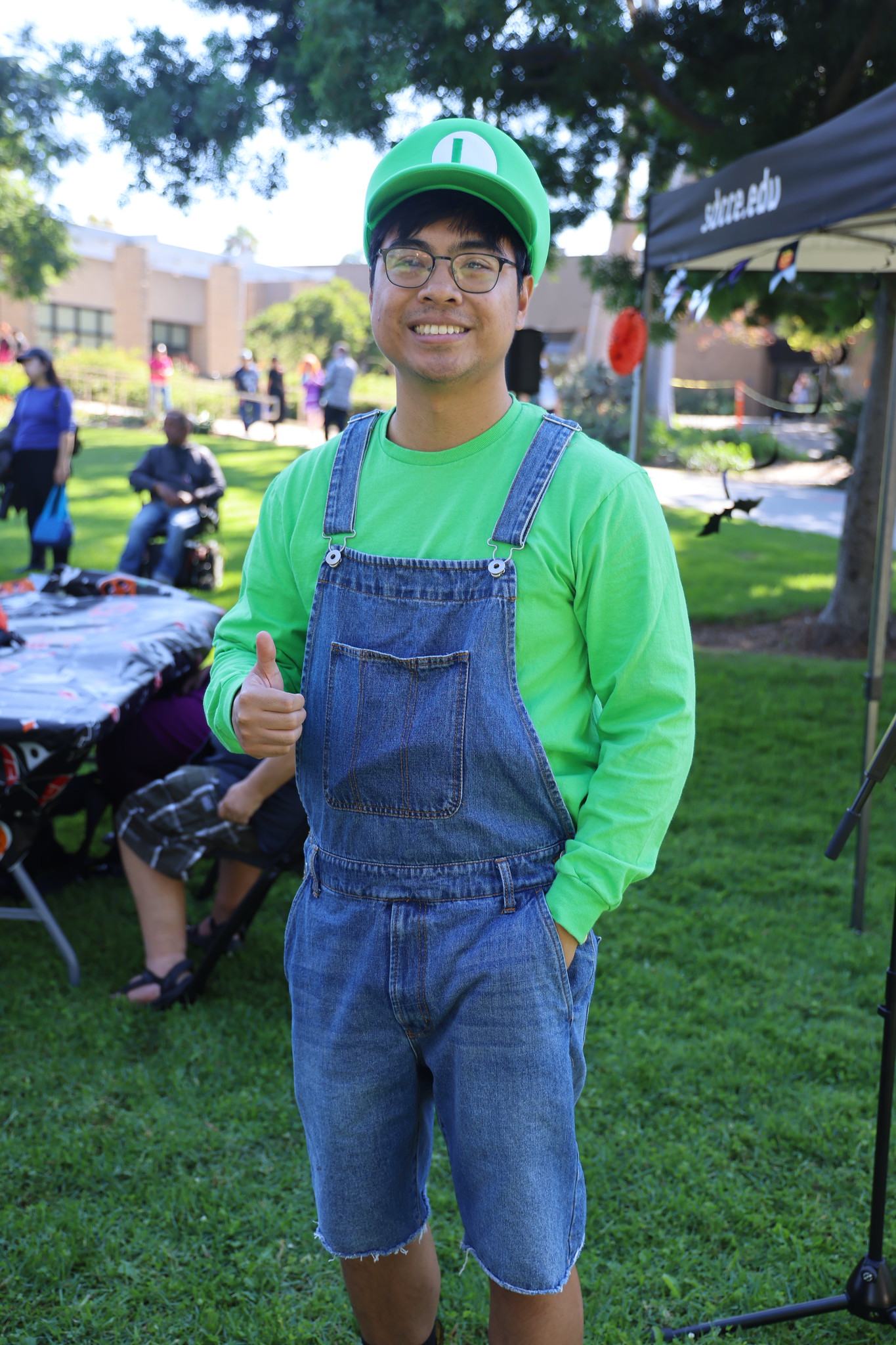 
A man in blue jean overalls and a green shirt and hat dressed as Luigi.
