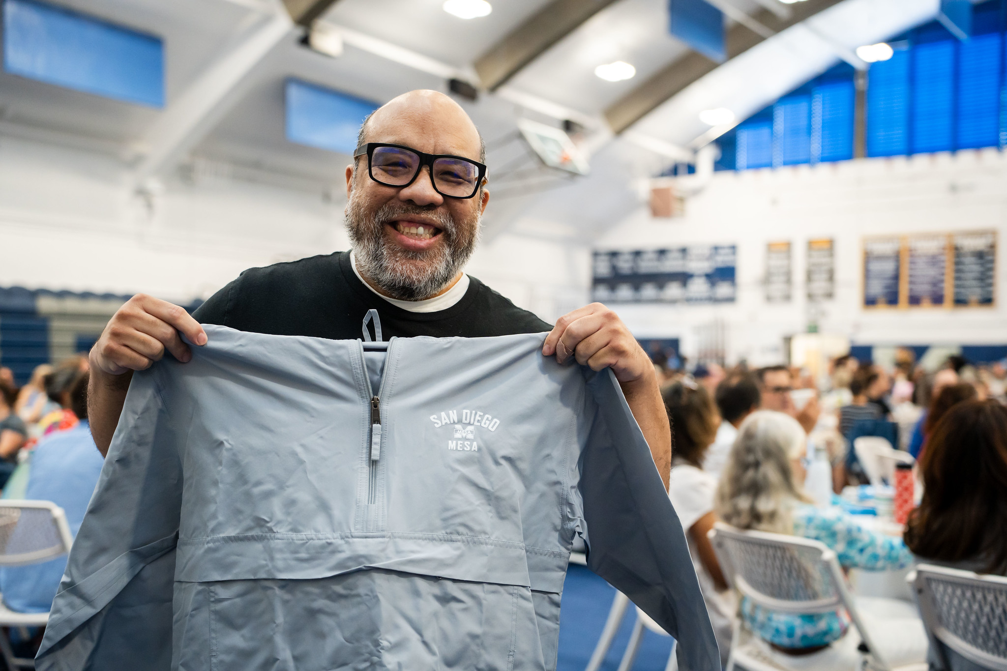 
A man holds up a light gray Mesa College windbreaker.
