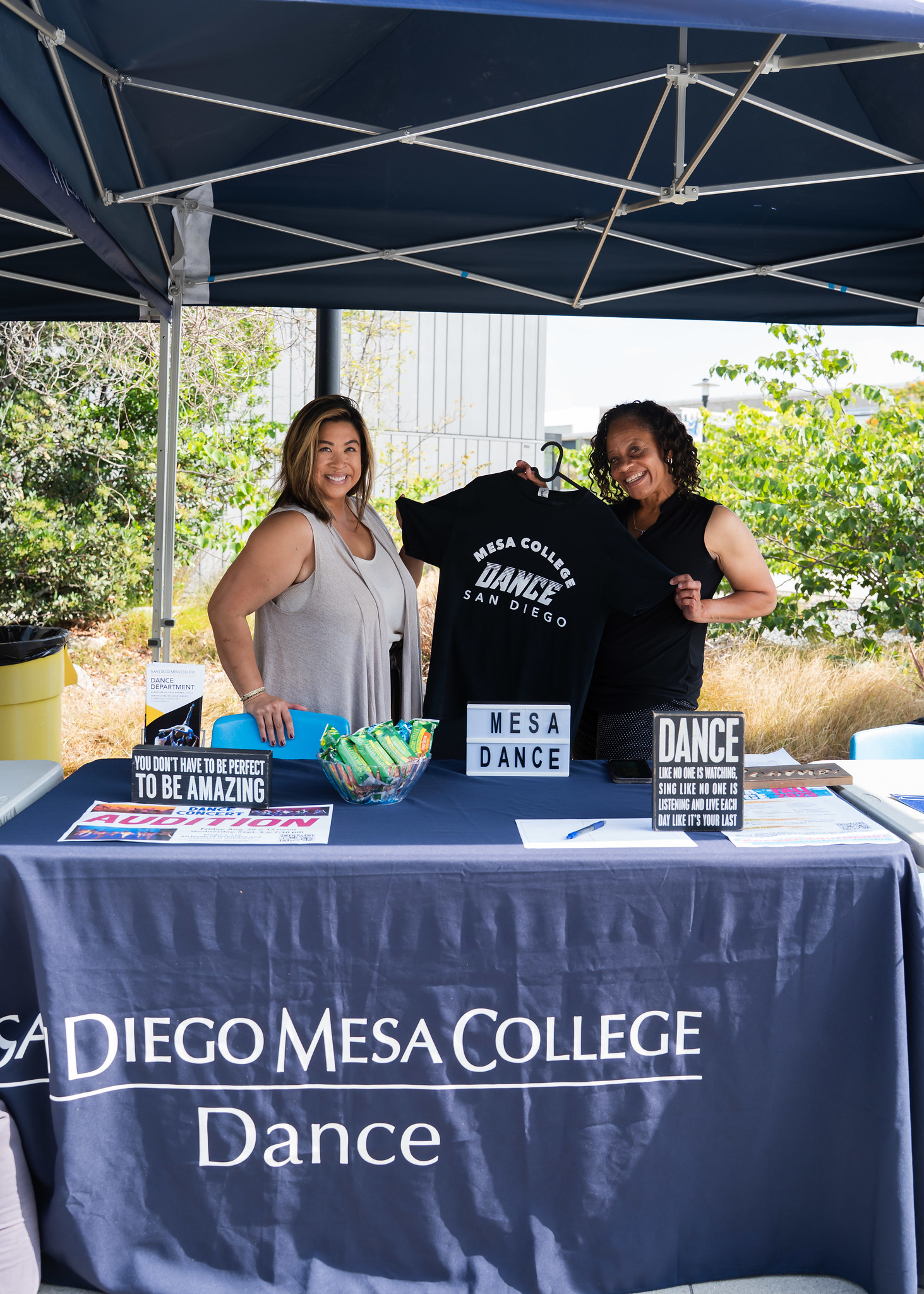 
Two women working at the Mesa College Dance welcome tent. One is holding up a black Mesa Dance t-shirt.
