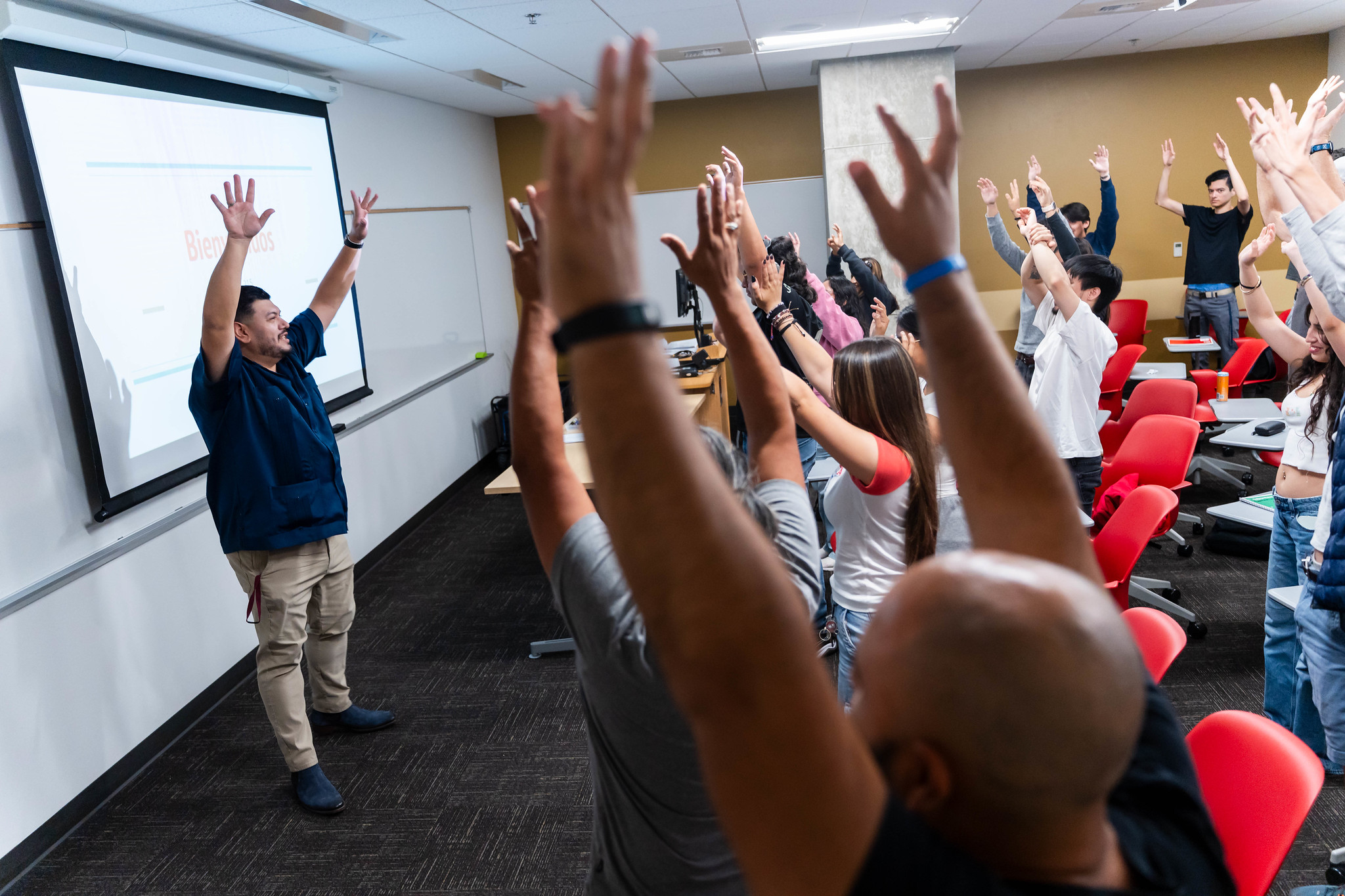 
People in a classroom standing with their arms raised over their heads.
