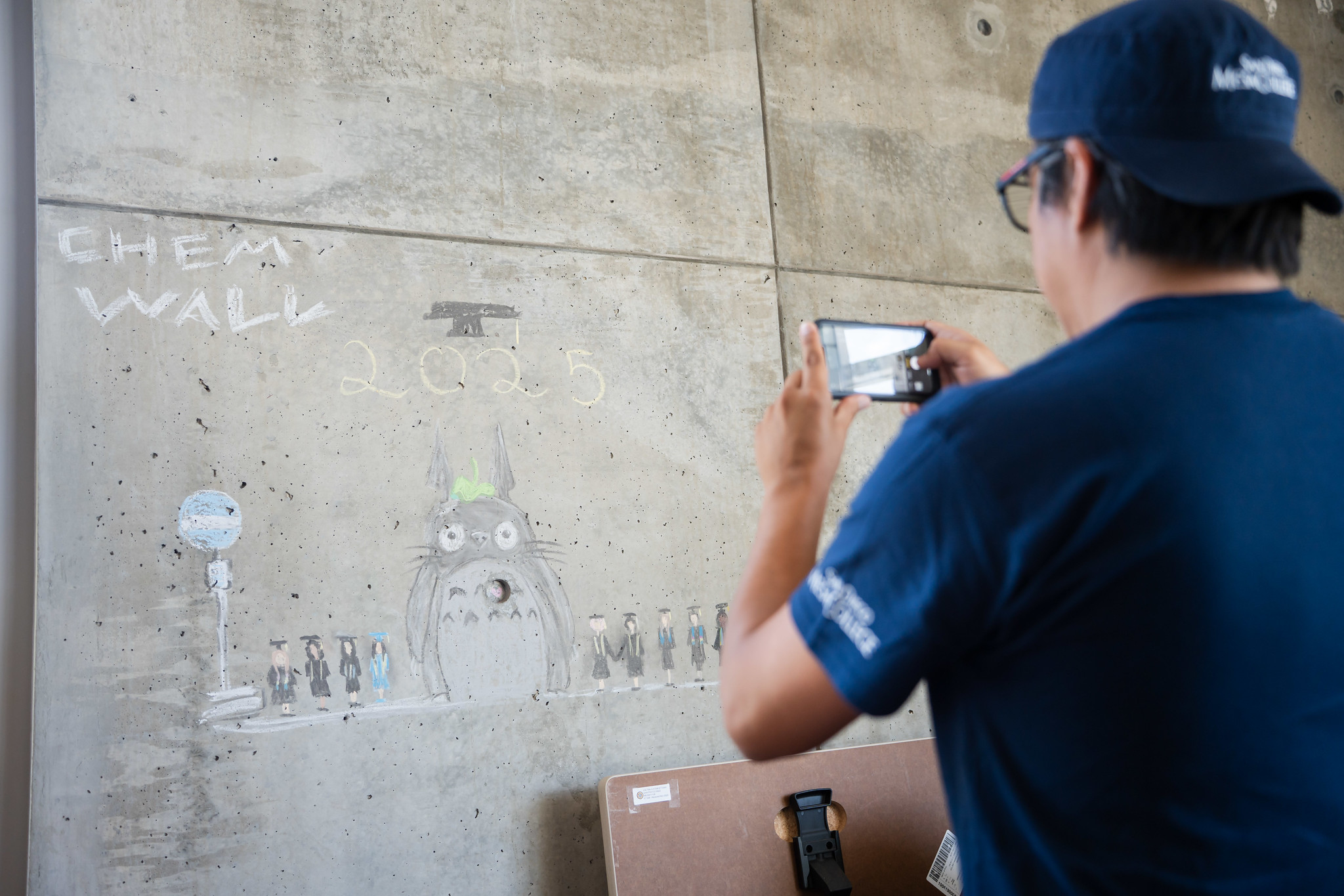 
A student uses his phone to take a picture of a chalk drawing on the wall of students in graduation caps and gowns
