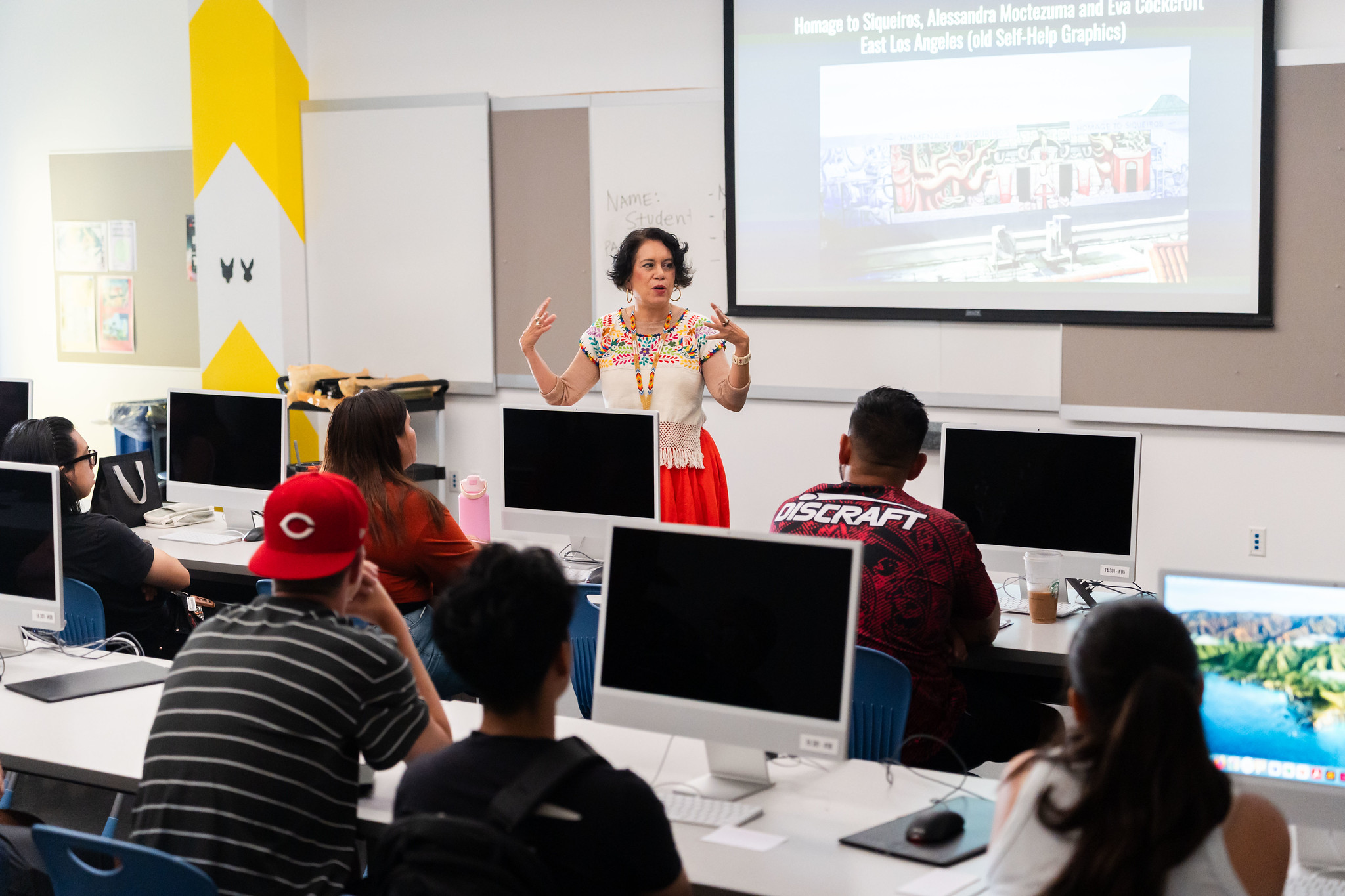 
An instructor talks to students seated at computer stations in class.
