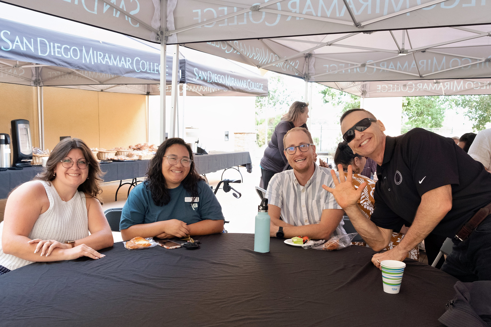 
Four people at a table. One is waving to the camera.
