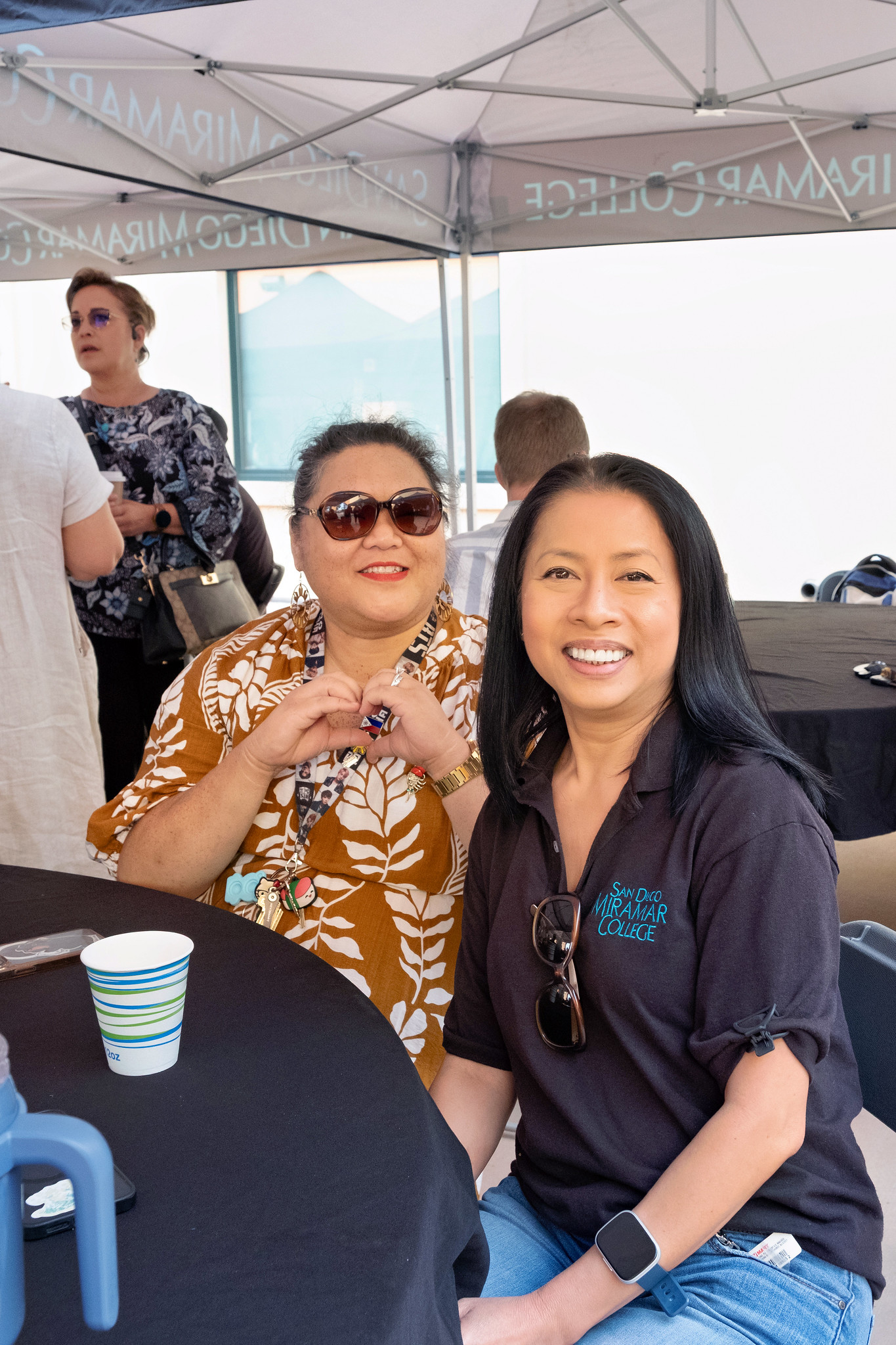 
Two ladies sitting at a table. One is making a heart with her hands.
