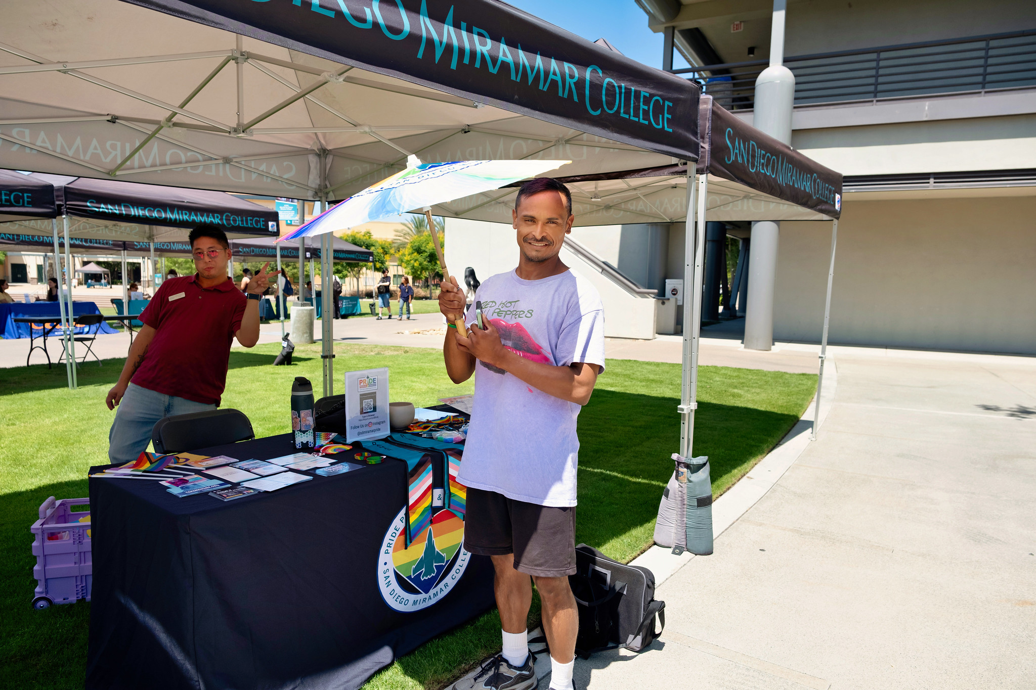 
A man holds up a rainbow umbrella while another man gives a peace sign at the Pride Center welcome booth.
