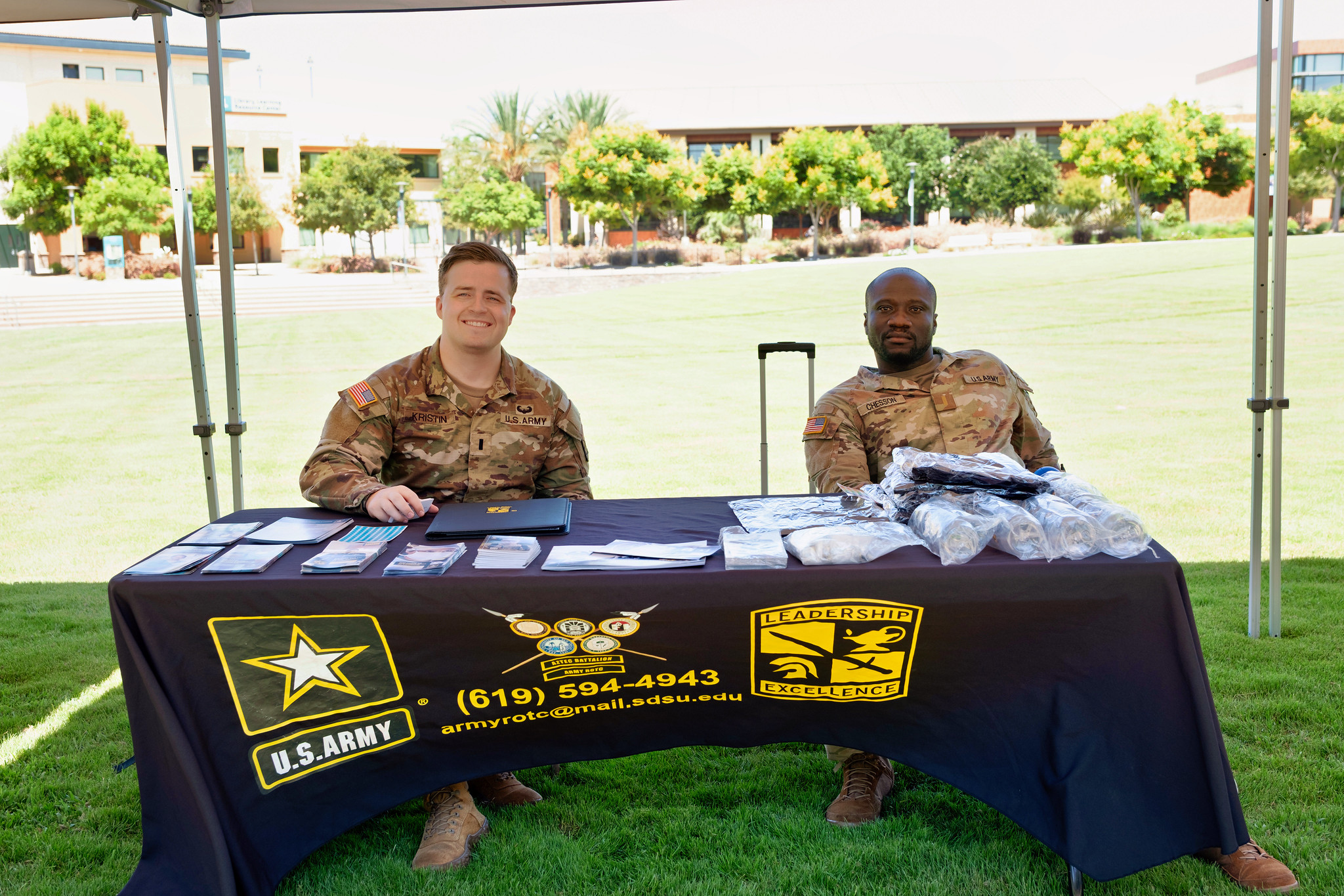 
Two men wearing camouflage sit at a table for military recruitment.
