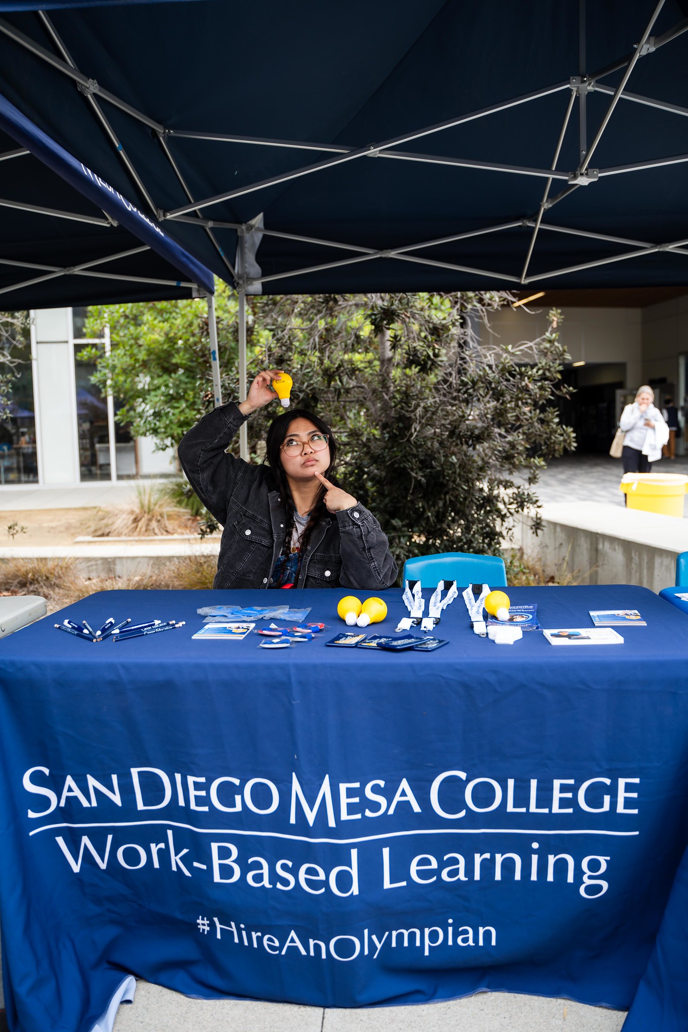 
A lady at a Mesa College booth holds a yellow lightbulb stress ball over her head.
