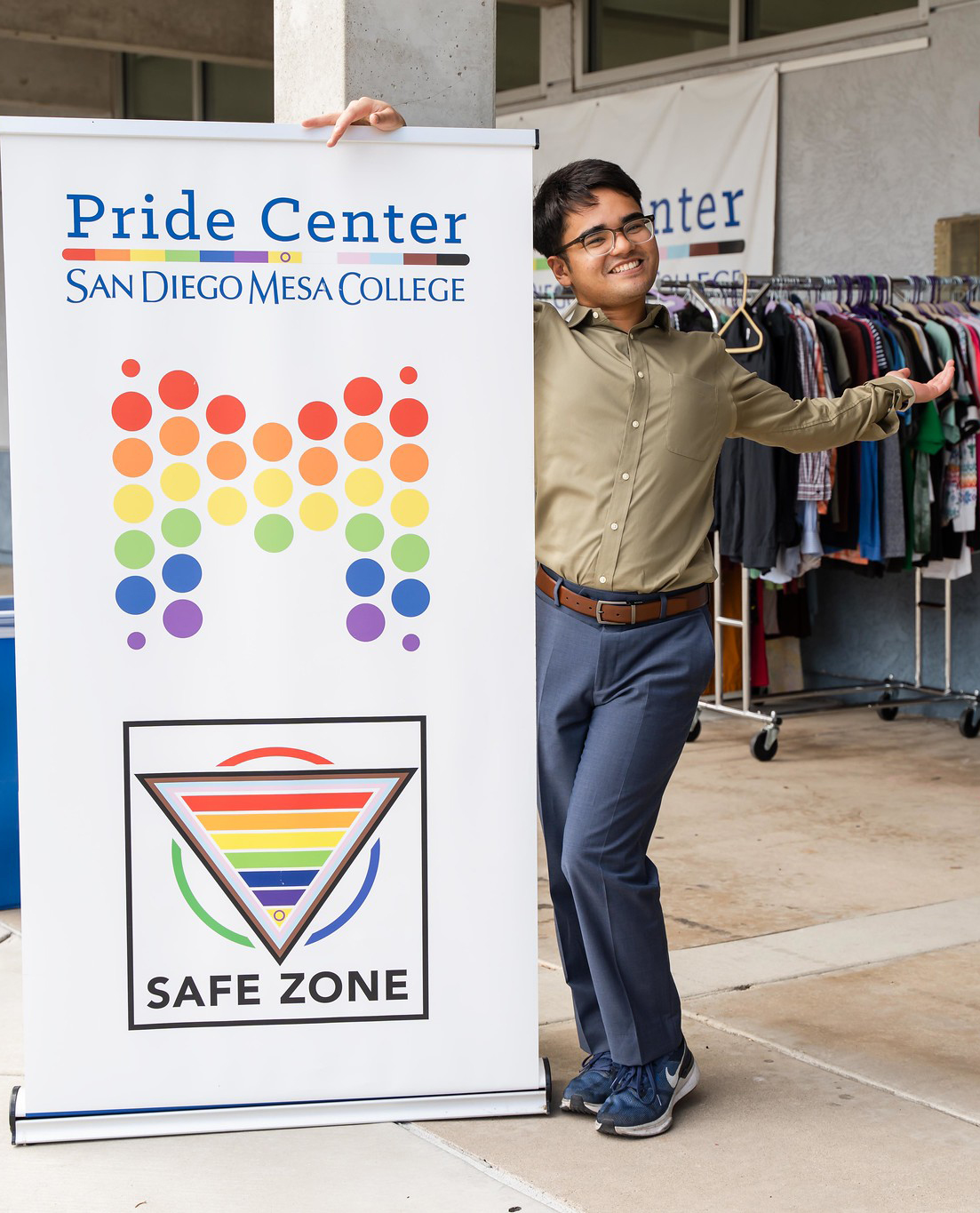 
A student standing next to a Mesa College Pride Center banner sign gestures toward the door of the pride center.
