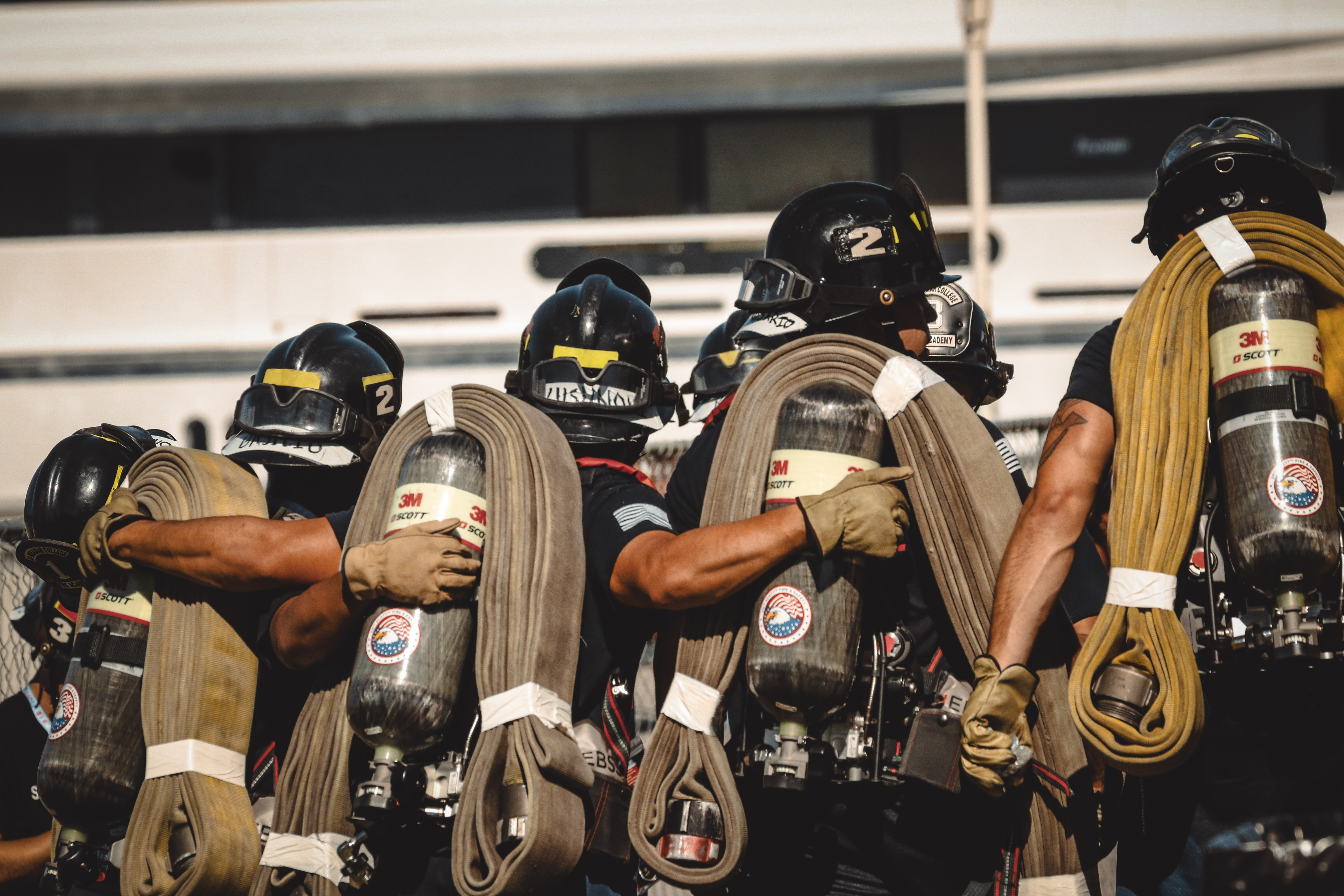
The back of five cadets wearing fire helmets and their gear put their arm around the cadet standing next to them.
