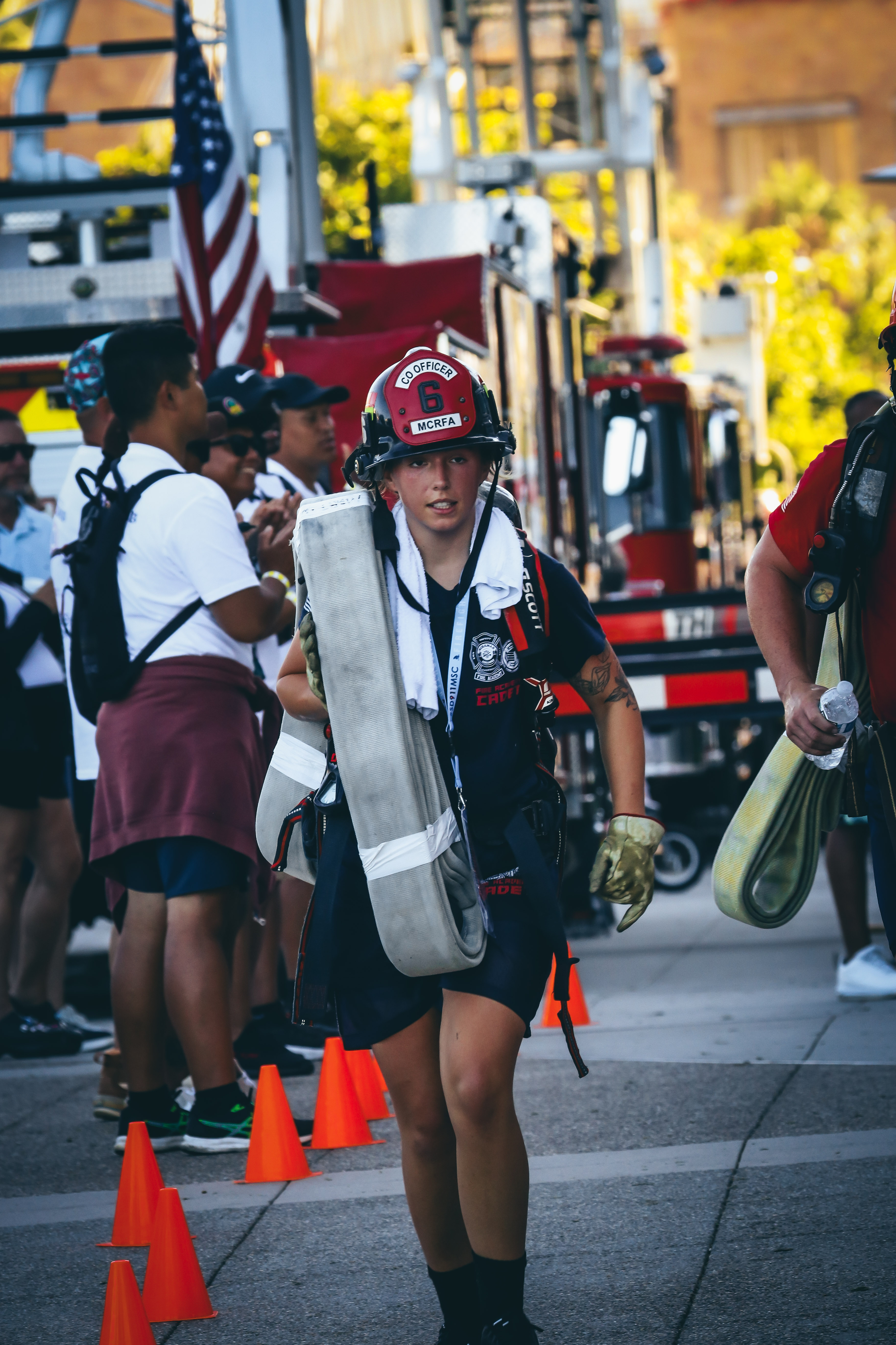 
A cadet at the event walking with a red fire helmet and fire gear and a fire hose.
