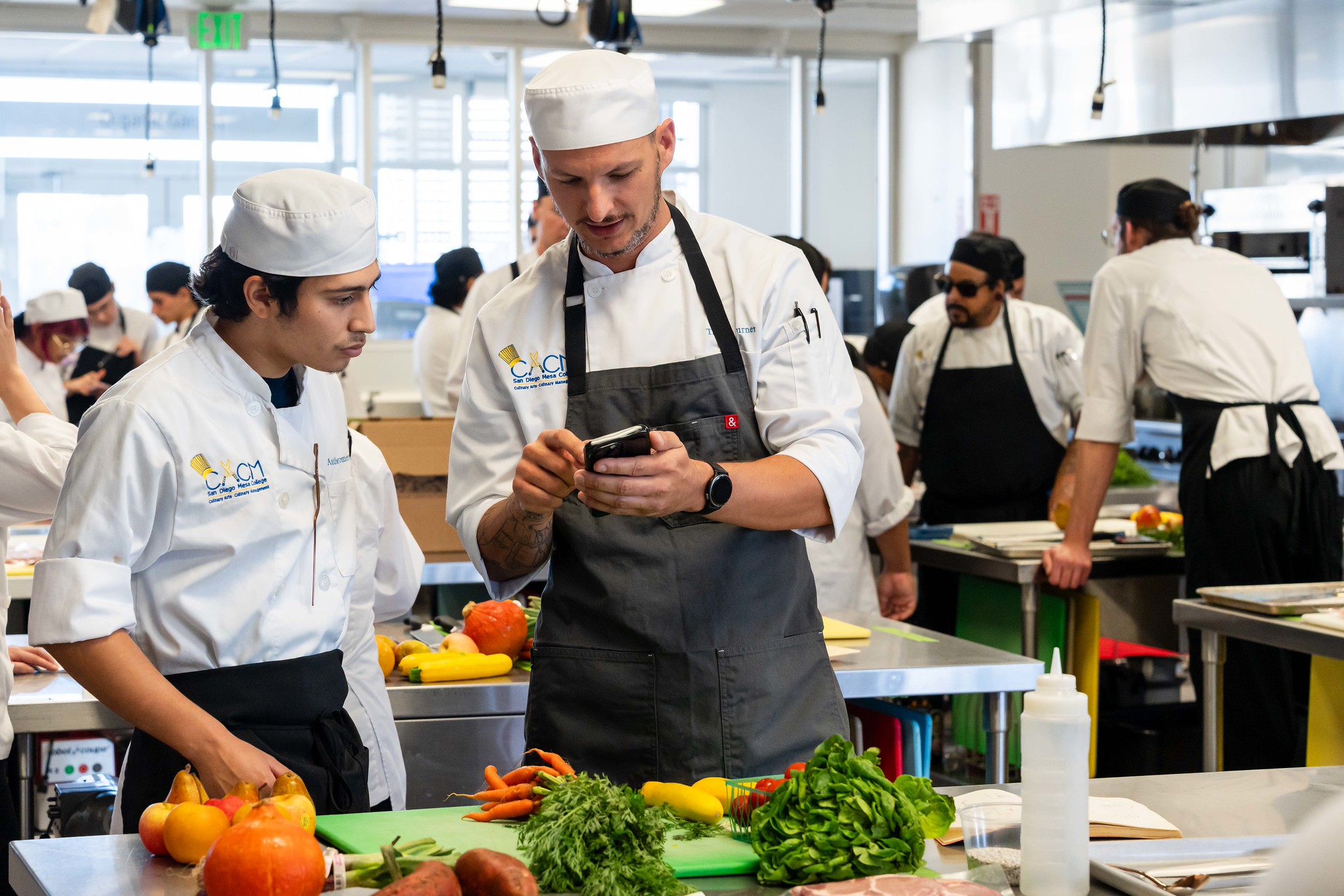 
Two chefs in the meal prep area stand in front of cutting boards full of vegetables. 
