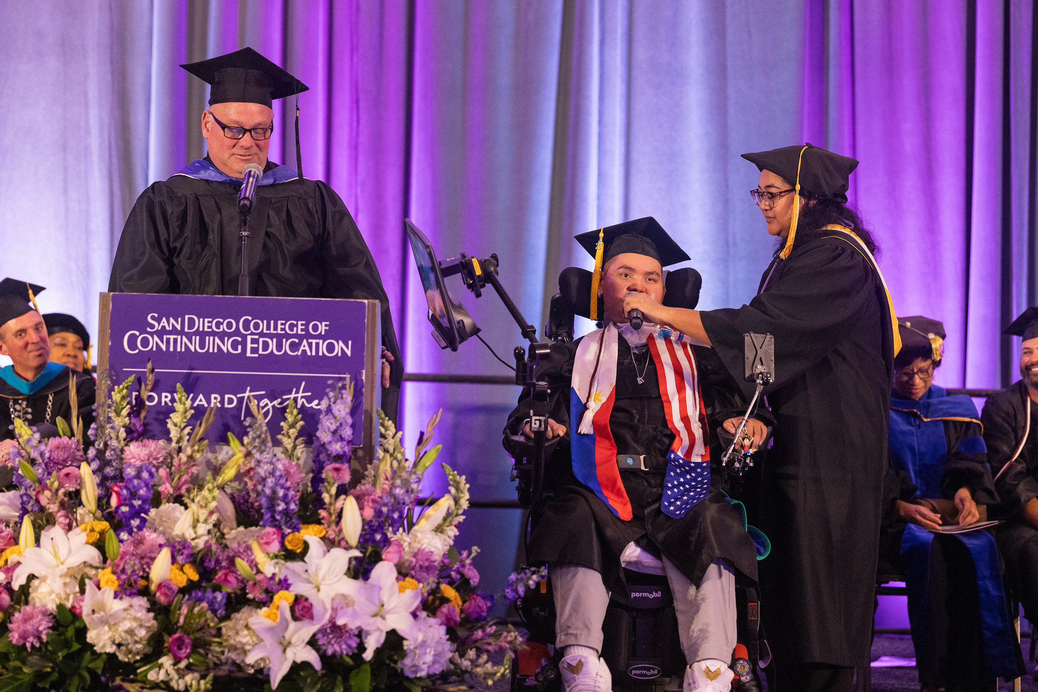 A speaker at a podium and a student using a wheelchair speak to the audience.