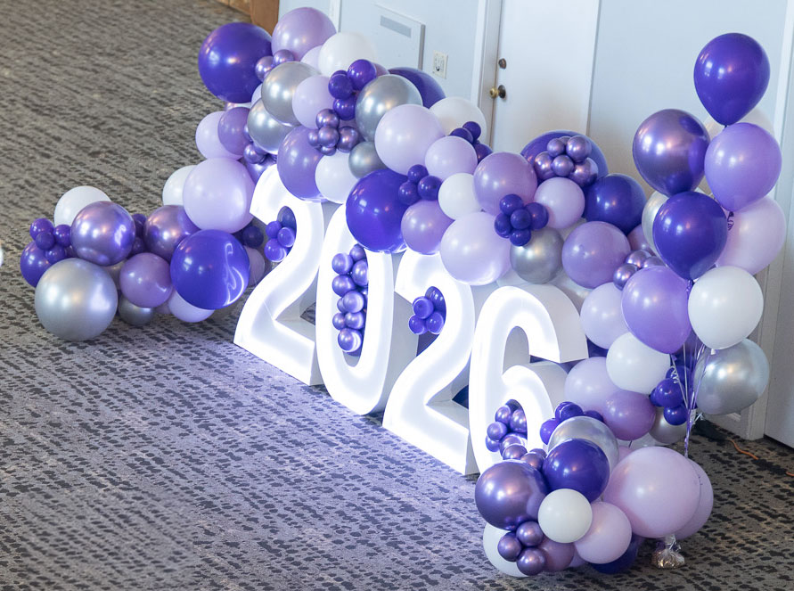 Large white lit up 2026 numbers surrounded by white and purple balloons for graduates to take photos around.