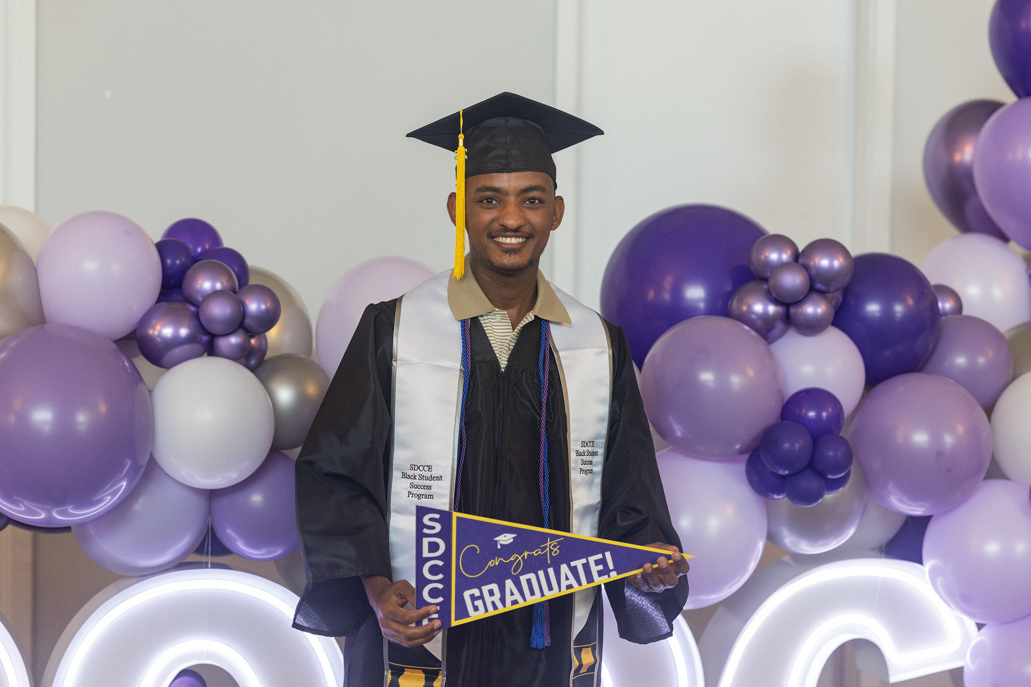 A graduate holds a purple graduate banner and stands in front of a lit up 2026 and balloons.