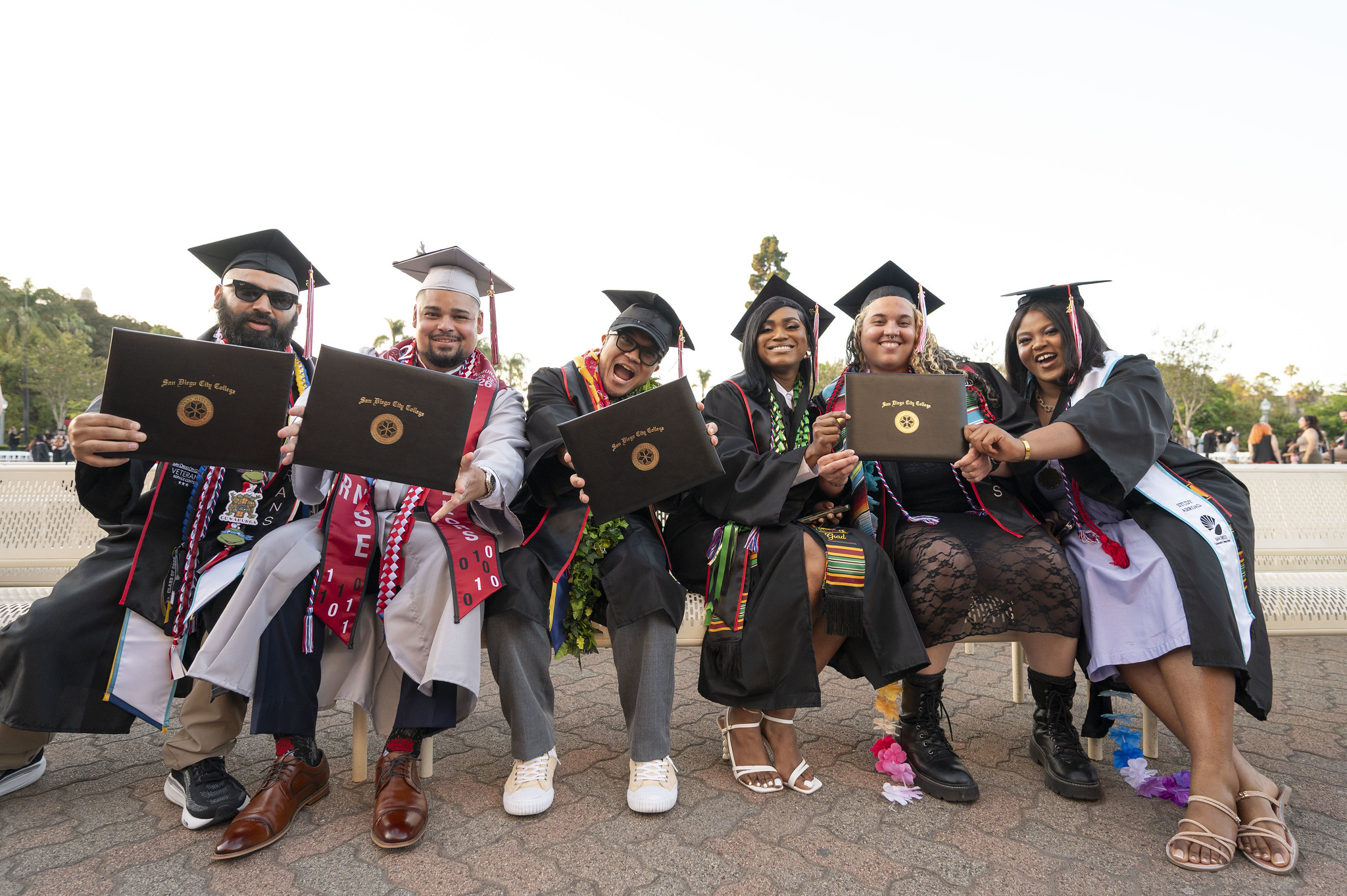 Six students in graduation caps and gowns hold up their degrees.&nbsp;