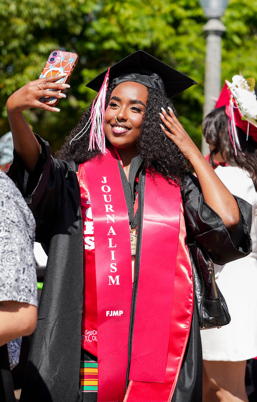 A graduate in a red journalism stole fixes her hair as she takes a selfie.&nbsp;