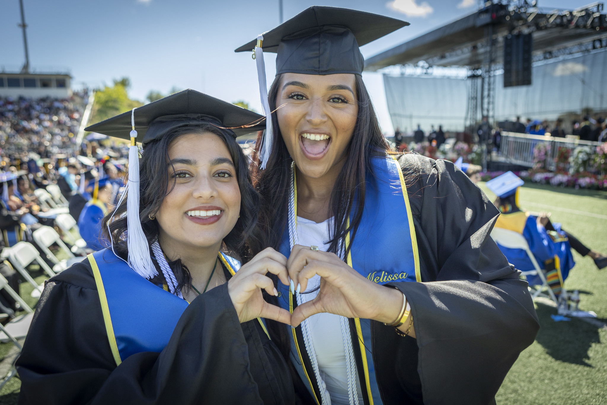Two graduates in caps and gowns put their hands together to make a heart.