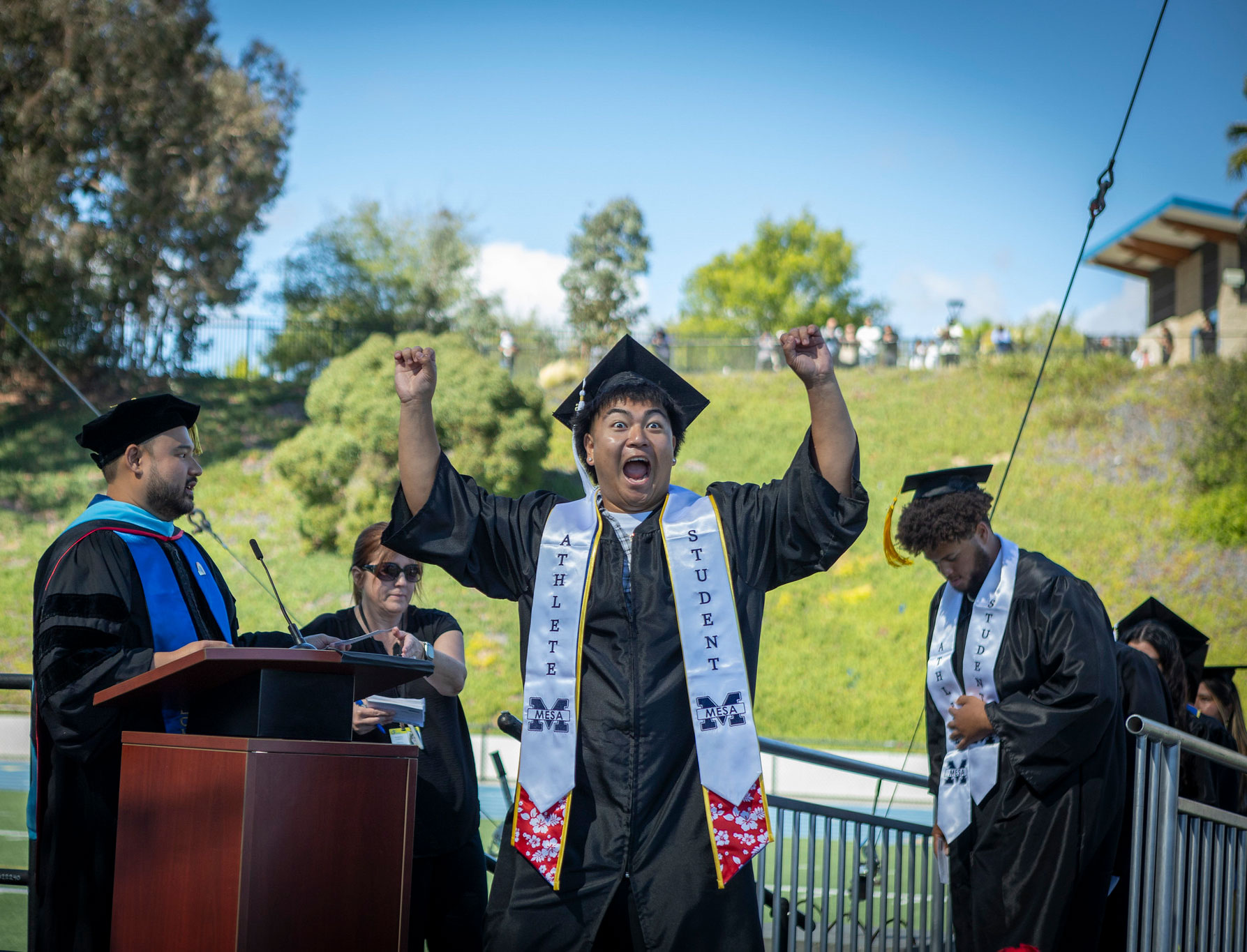A grad faces the audience with both hands in the air as he cheers on stage.