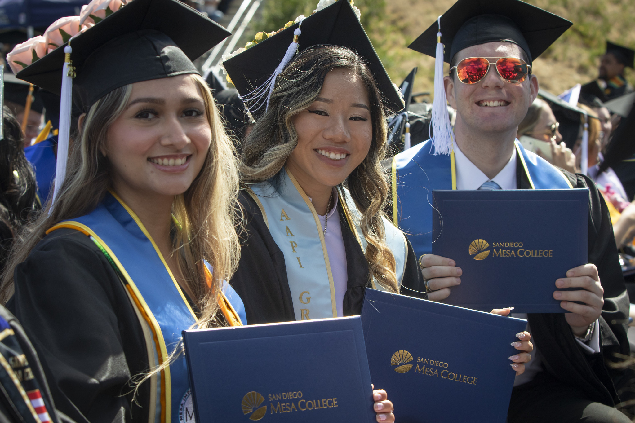 Three grads seated in the audience hold up their degrees.&nbsp;
