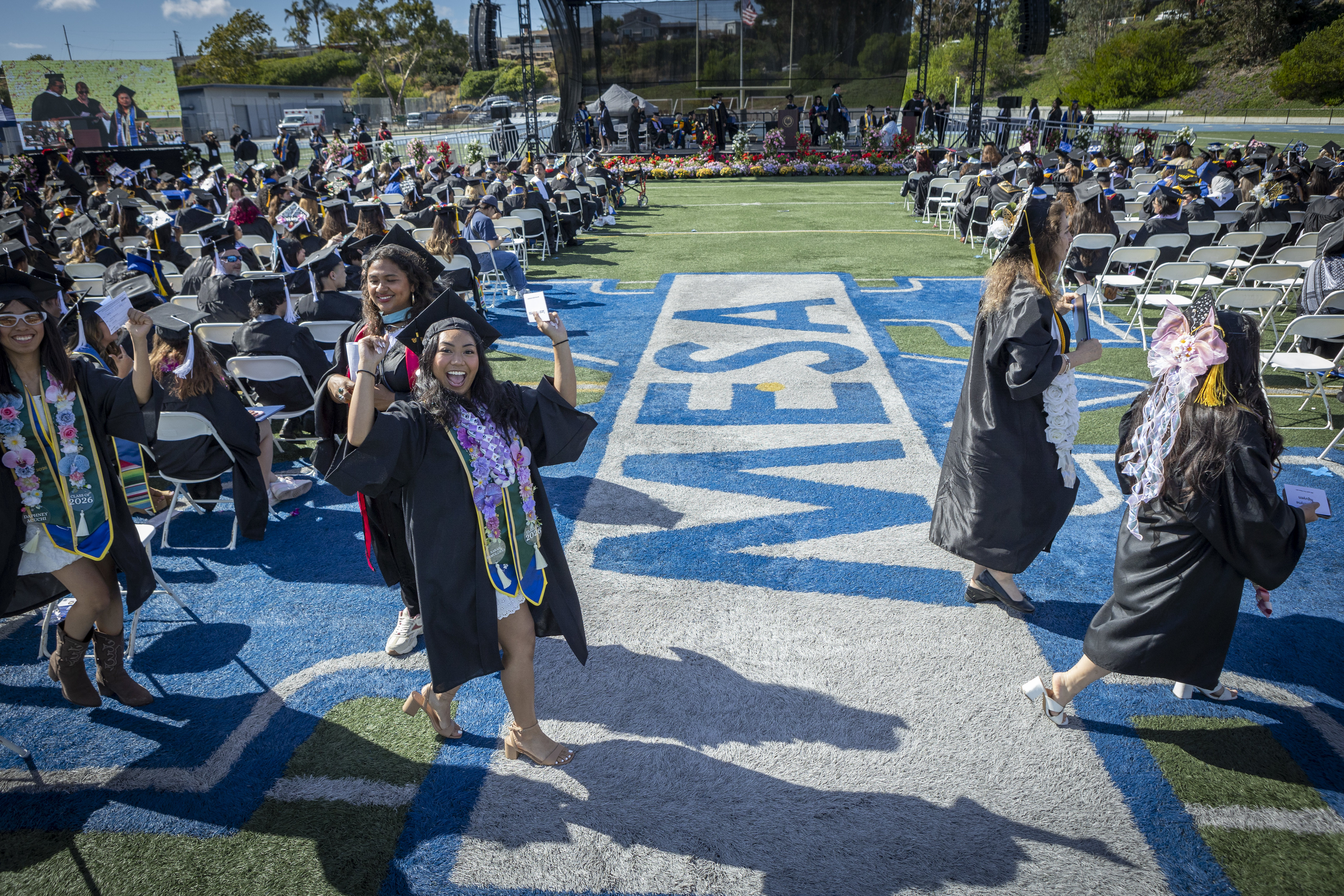 A student cheers in front of the blue and white Mesa logo painted on the grass.