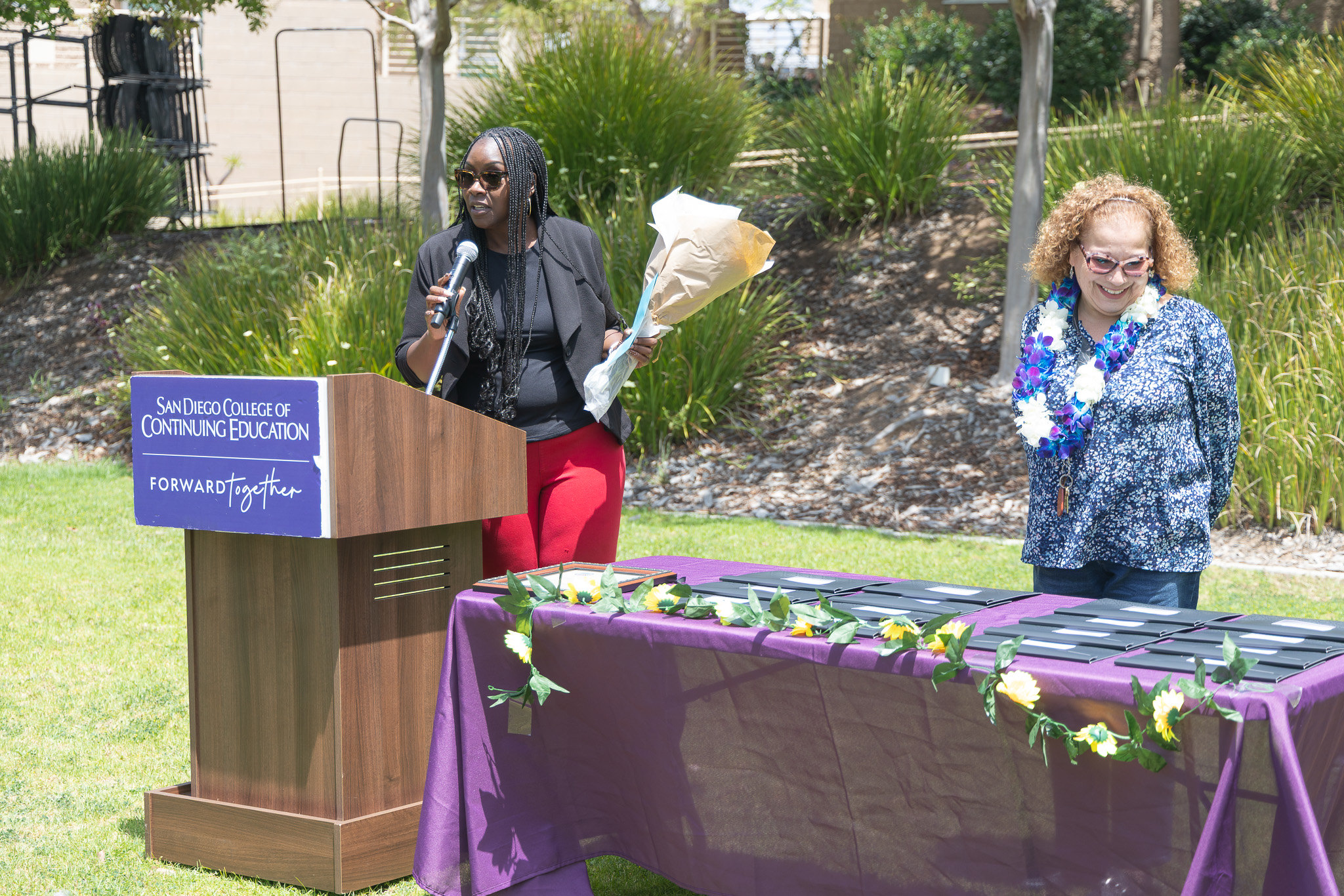 A presenter holds a bouquet of flowers while another presenter looks over the table of certificates for employees' service.