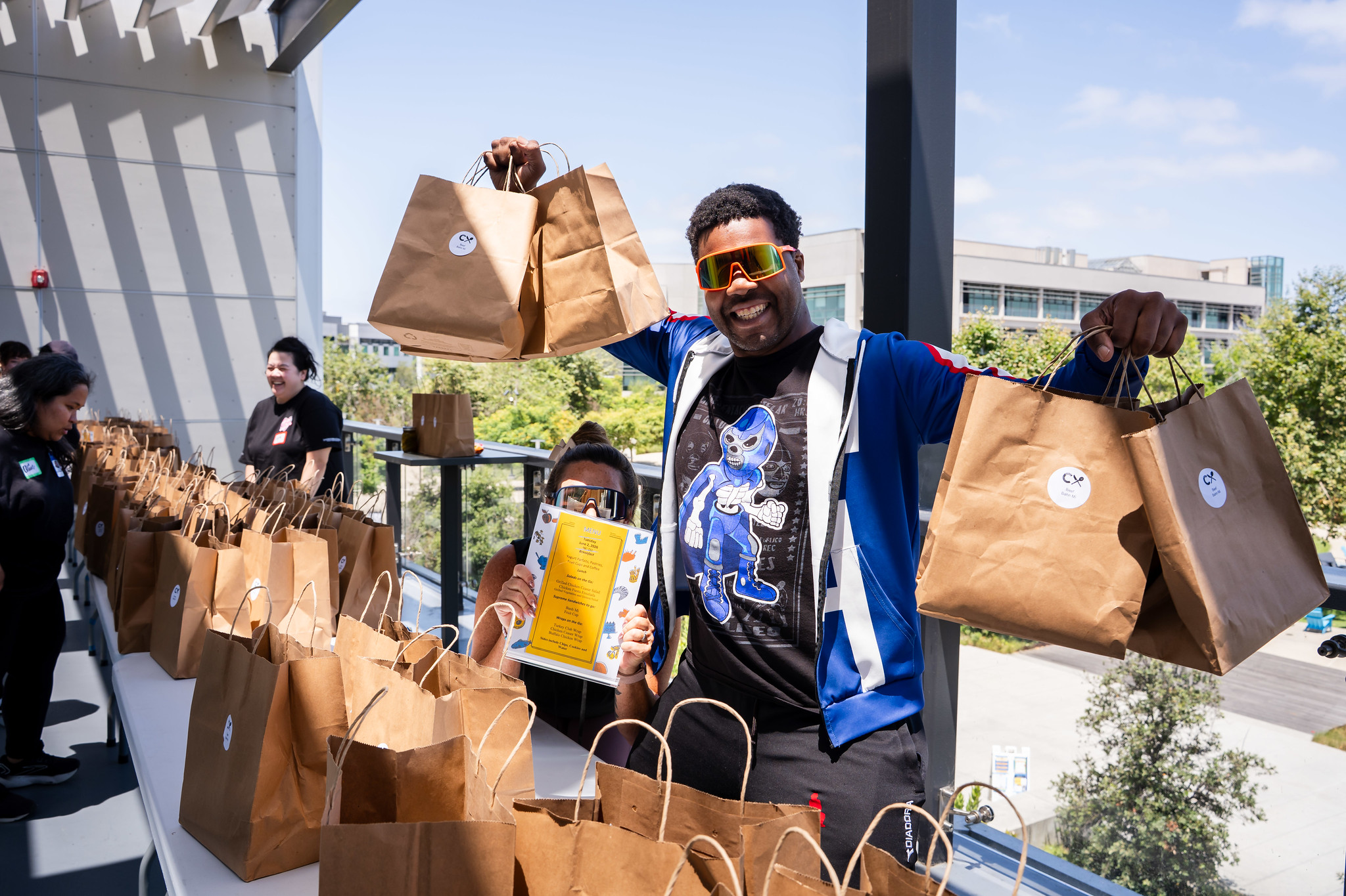 An employee stands behind a table of dozens of bags, holding up four goodie bags being given away to employees.&nbsp;